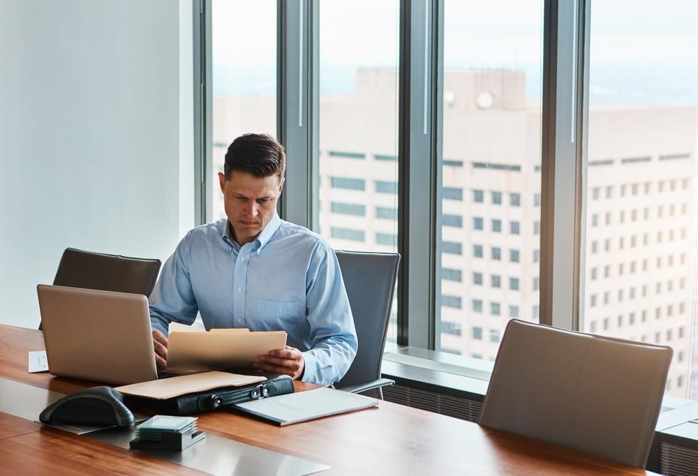Man reading documents at an office conference table with a laptop, city buildings visible through the windows