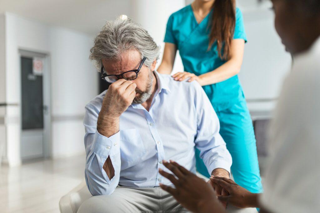 An older man sitting in a medical setting with his head lowered and fingers pinching the bridge of his nose in distress, while a healthcare worker in teal scrubs gently places a hand on his shoulder and another person in the foreground speaks to him with hands slightly extended.