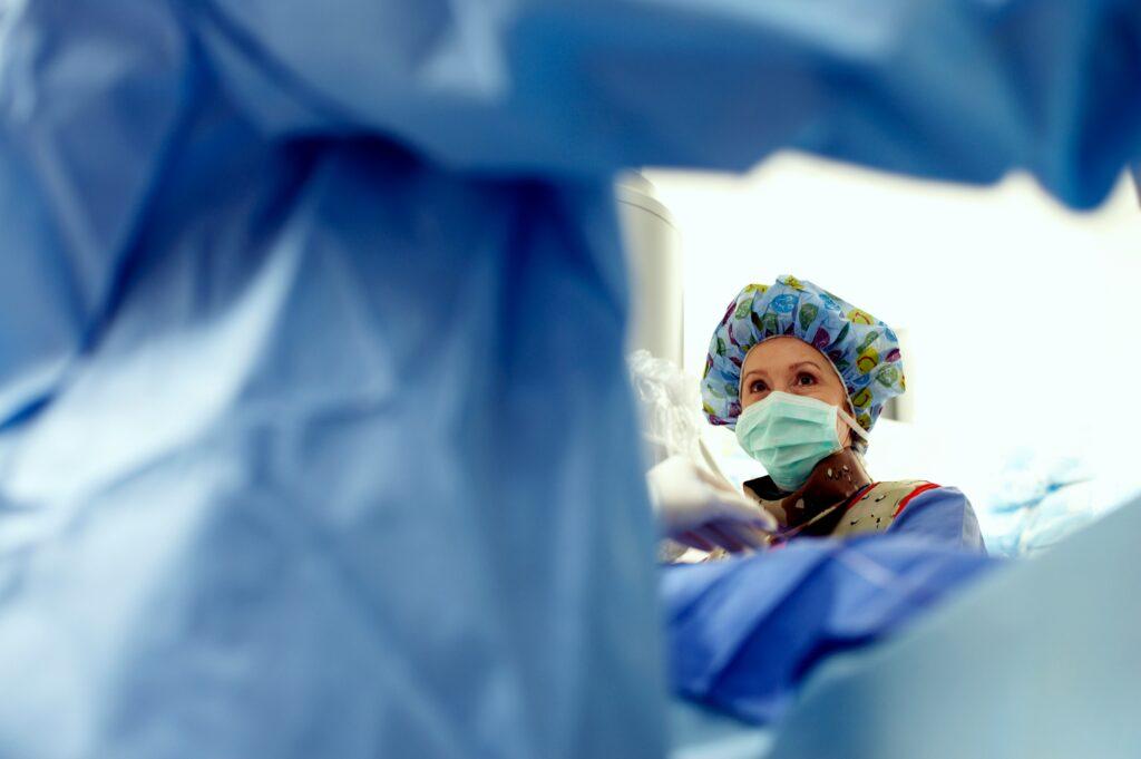 A medical professional wearing a surgical mask, patterned scrub cap, and protective gown looks down attentively during a procedure, seen from the patient’s perspective through blurred blue surgical drapes, with bright clinical lighting and a focused operating room atmosphere.