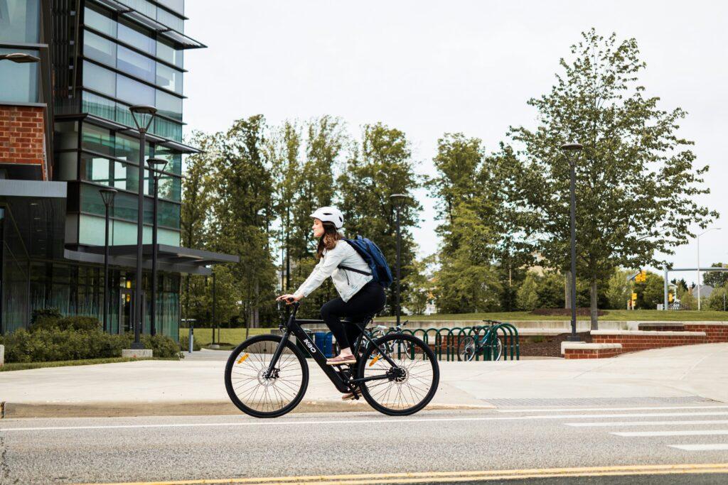 A woman wearing a helmet and backpack riding a bicycle along a city street near a modern building and a row of trees in the background.