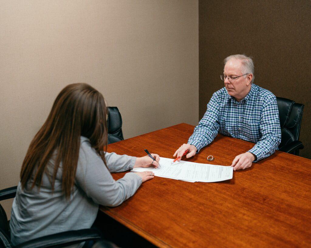 A woman seated at a conference table signs a document with a pen while an older man across from her points to specific sections on the paperwork, both focused on the forms spread out on a wooden table in a quiet office meeting room.