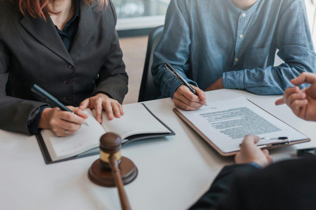 Two people seated at a table signing and reviewing legal documents, one writing in a notebook and the other signing a printed agreement on a clipboard, with a judge’s gavel resting on the table, suggesting a formal legal consultation or contract signing meeting.