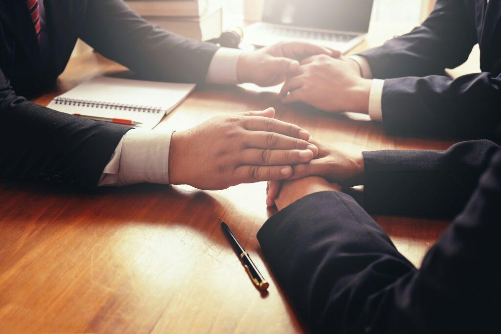 Three people in business attire sit around a wooden table, with one person placing a reassuring hand over another’s hand while documents, a notebook, and pens rest nearby, conveying support, empathy, and a serious professional discussion.