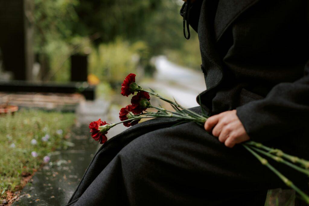 A person dressed in dark clothing sits on a park bench holding a small bouquet of red carnations in one hand, with a blurred cemetery or memorial setting in the background, green foliage around the path, and a quiet, somber atmosphere suggesting remembrance or mourning.