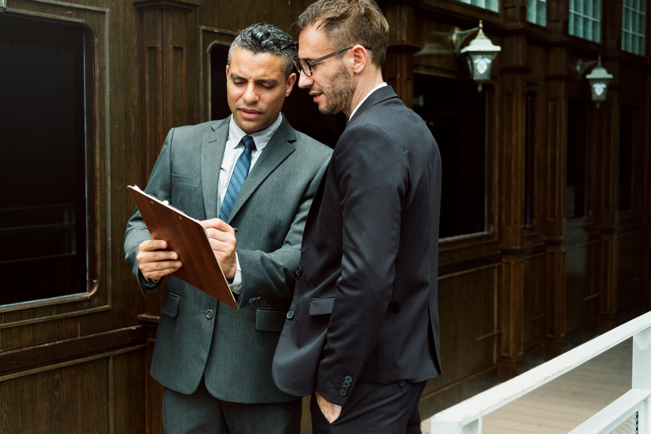 Two men in suits stand on a porch, examining a clipboard together.