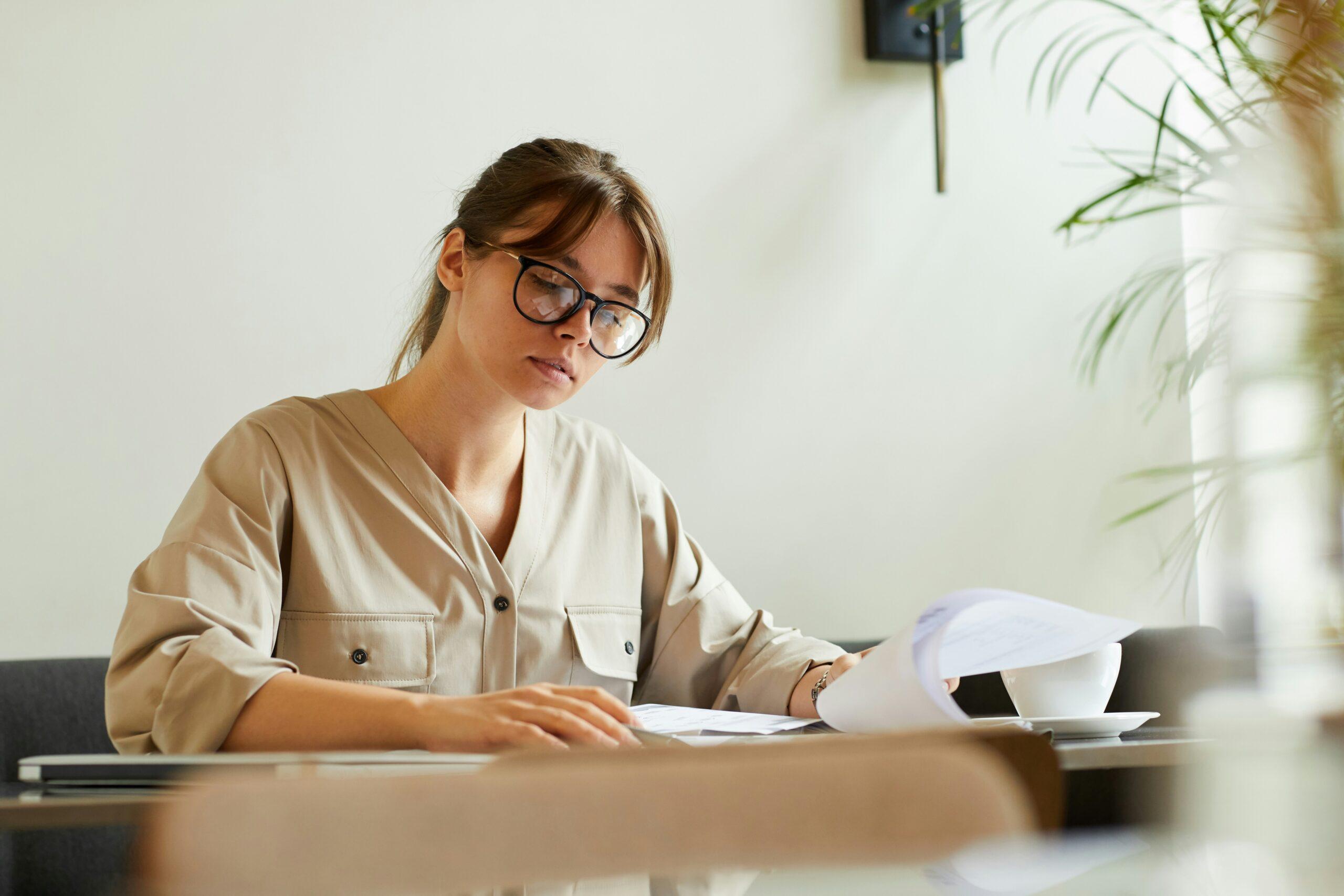 A woman wearing glasses sits at a table, reviewing papers with a focused expression.