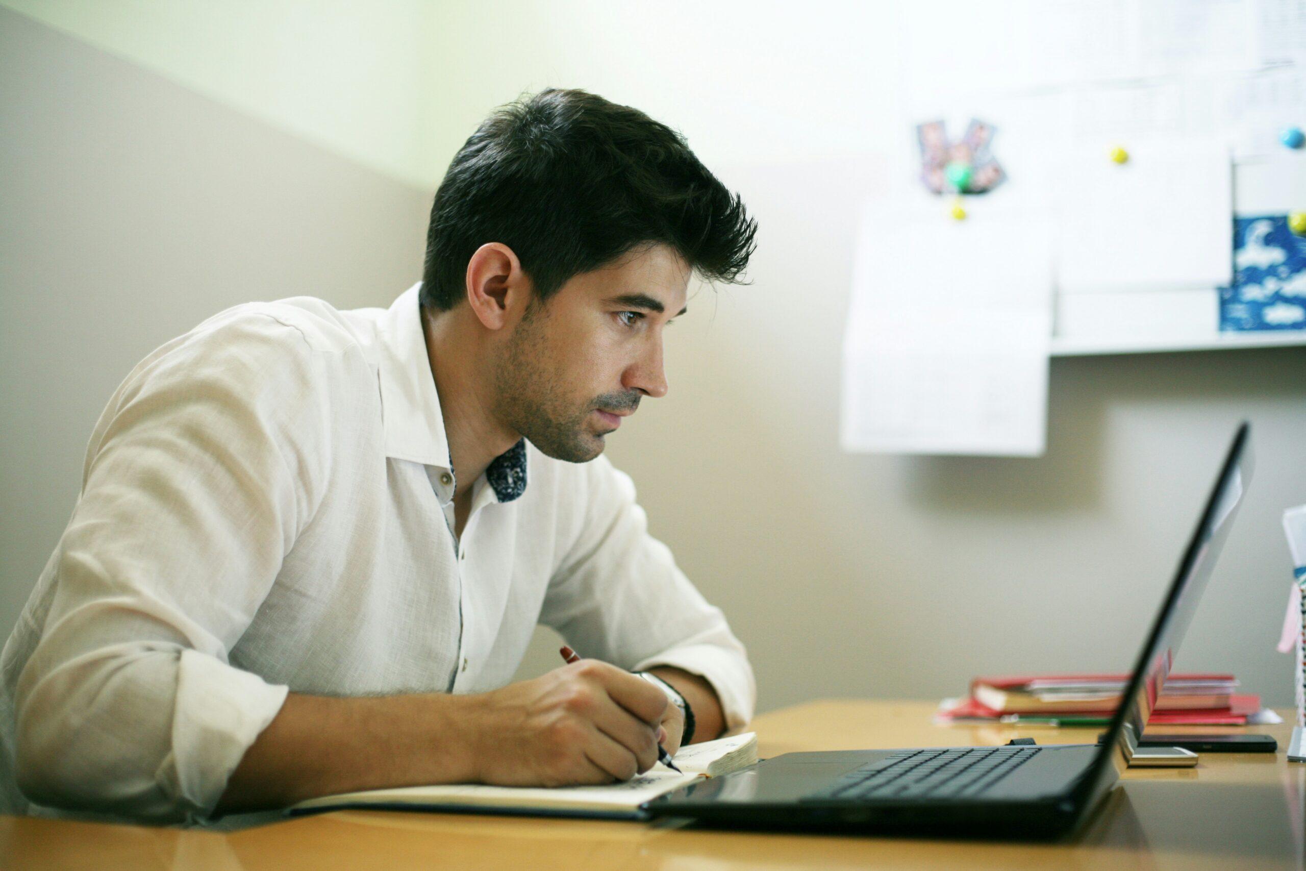 A man sitting at a desk, focused on his laptop, with papers and a coffee cup nearby.