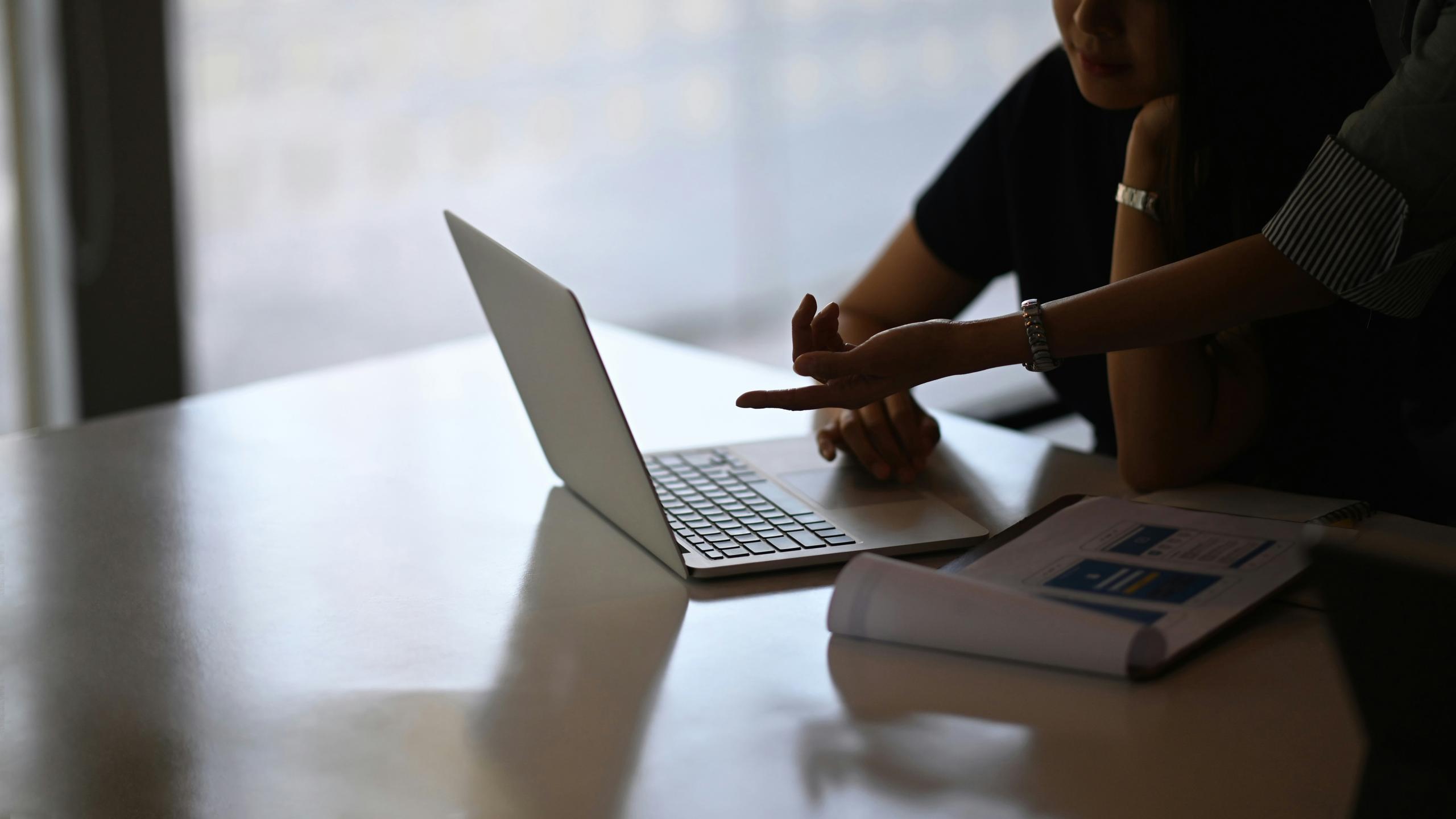 A woman points at a laptop on a table, highlighting its features or content during a discussion.