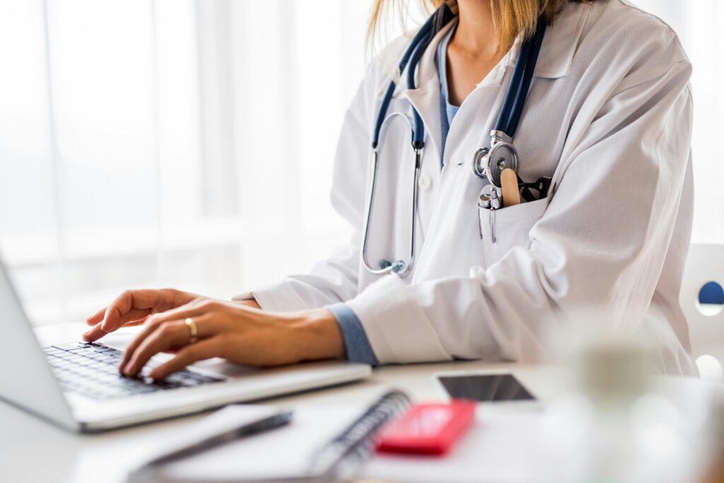 A woman in a doctor's coat types on her laptop, focused on her work in a medical setting.