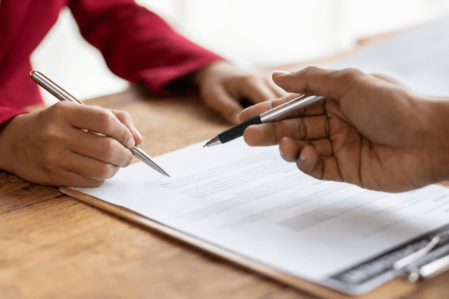 Close-up of two people reviewing and signing documents at a wooden table with pens in hand and a blurred office background