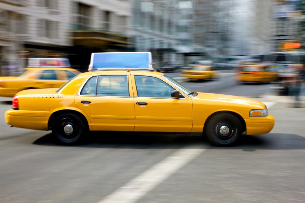 A yellow taxi cab driving along a busy city street, surrounded by tall buildings and pedestrians.