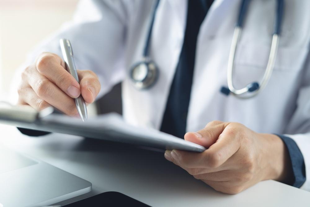 A doctor in a white coat writes notes on a clipboard in a clinical setting, focused on patient information.