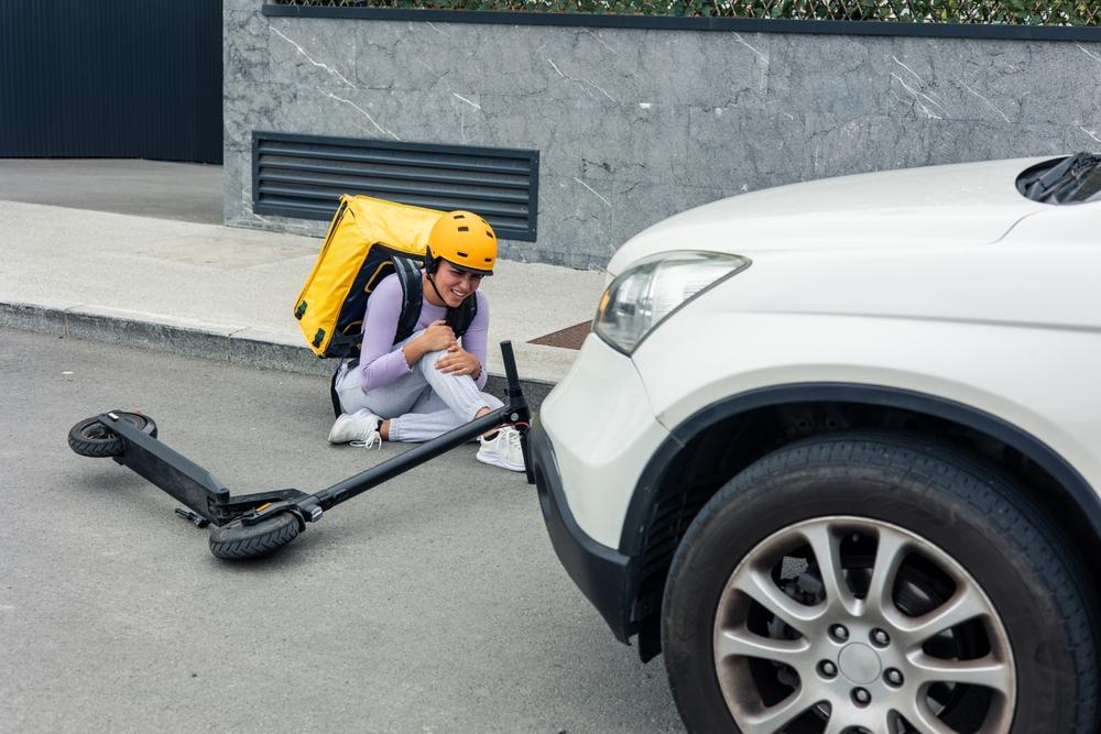Injured delivery rider with a helmet and backpack sitting on the road holding their knee beside a fallen electric scooter and a car
