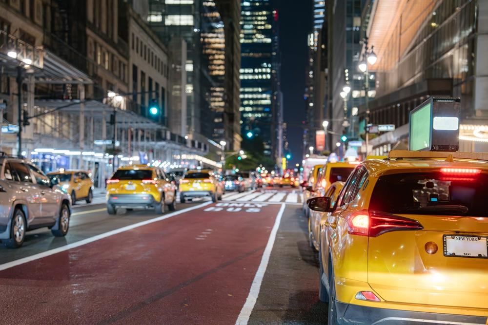 A bustling city street at night, illuminated by streetlights, featuring cars and taxis in motion.