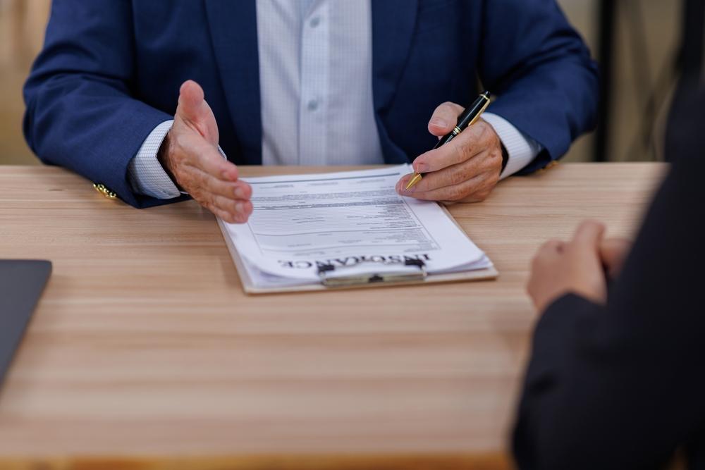 A man in a suit signs paperwork while seated across from a woman, both engaged in a professional discussion