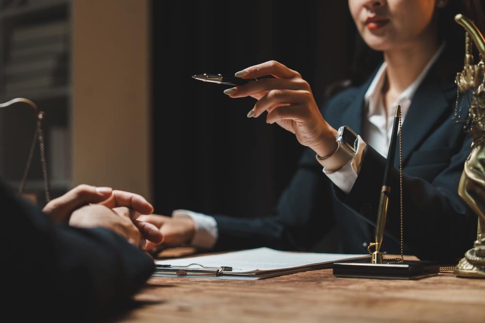 Woman in a suit handing a pen across a desk to a client with documents and scales of justice on the desk