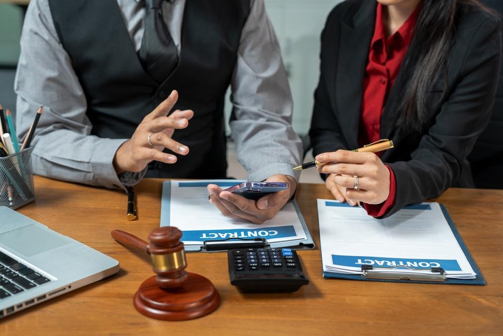 Two people at a desk with a gavel, calculator, and clipboards, text on documents: Contract