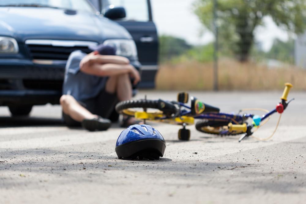 Man in a suit kneeling beside an injured cyclist lying on the ground next to a fallen bike and silver car
