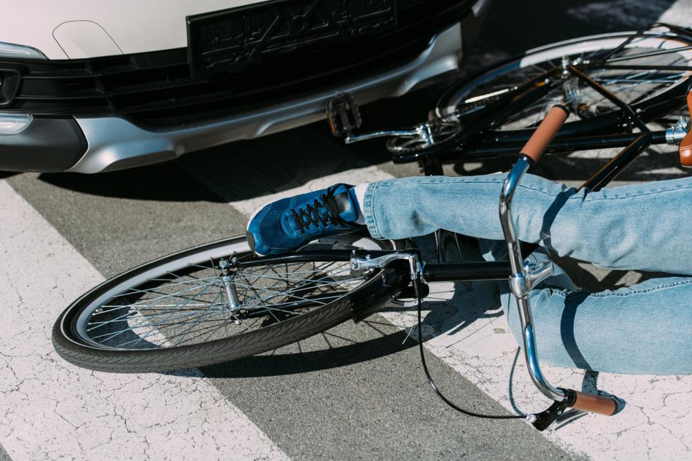 Cyclist in jeans lying on the ground beside a fallen bike in front of a silver car after a collision